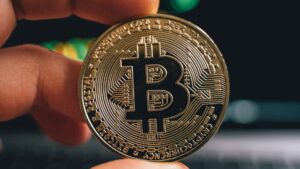 Mano sosteniendo una moneda de Bitcoin frente a una pantalla de trading de altcoins