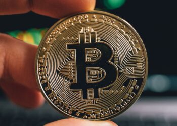 Mano sosteniendo una moneda de Bitcoin frente a una pantalla de trading de altcoins